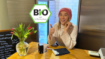 Male Müller hinter dem Tresen im Verkaufsladen, lachend und mit Daumen nach oben. Auf dem Tresen steht links ein Tulpenstrauß in einer bauchigen Vase. Oben im Vordergrund: das Biosiegel. Im Hintergrund: Die von innen beleuchteten, noch leeren Kühlschränke.